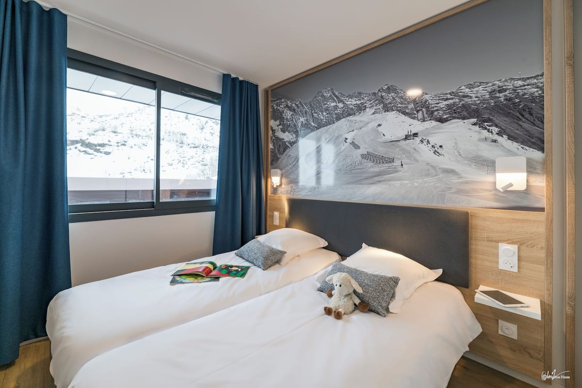 Quarto de hotel com duas camas, uma janela grande com cortinas, um quadro na parede retratando uma montanha nevada e uma mesa de cabeceira com uma luminária e um livro.