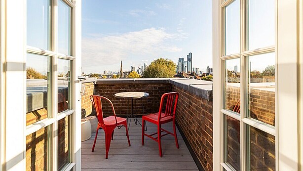 Eine Dachterrasse mit zwei roten Stühlen und einem kleinen runden Tisch, mit Blick auf eine Stadtansicht.