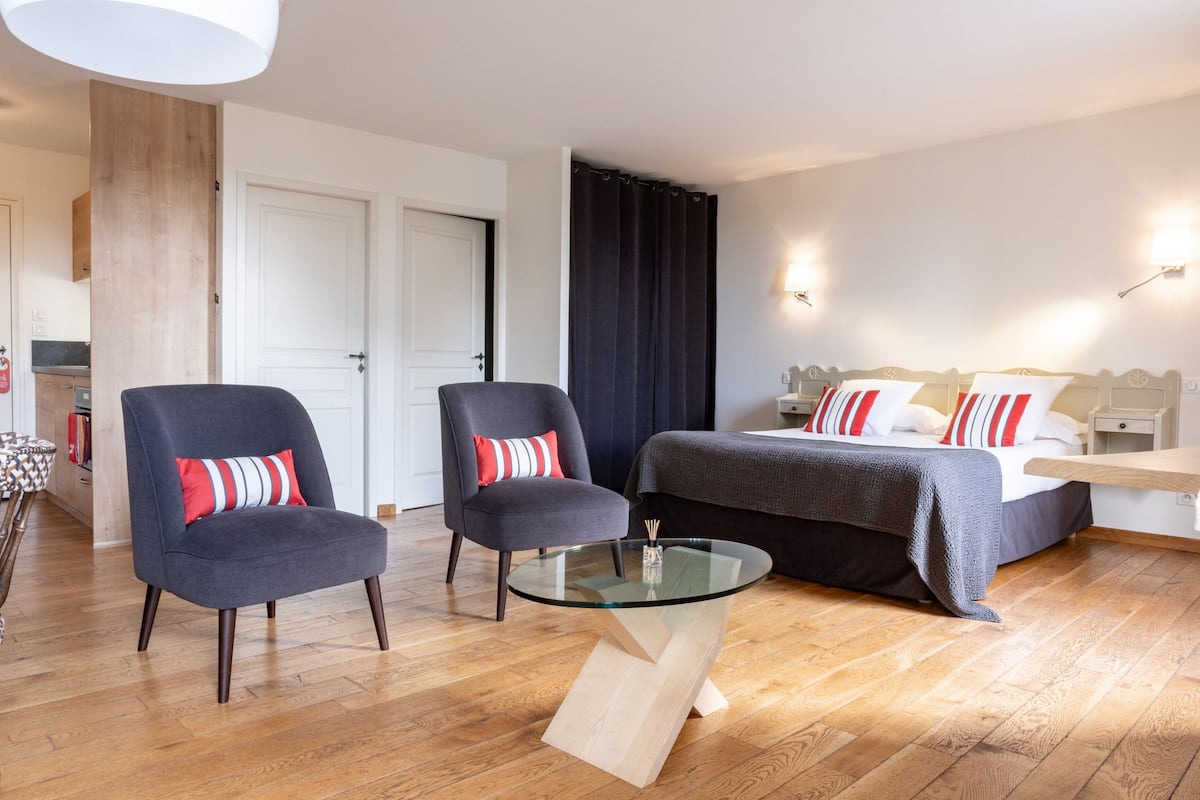 A modern hotel room with a bed, two armchairs, a glass coffee table, wooden flooring, and a white door.