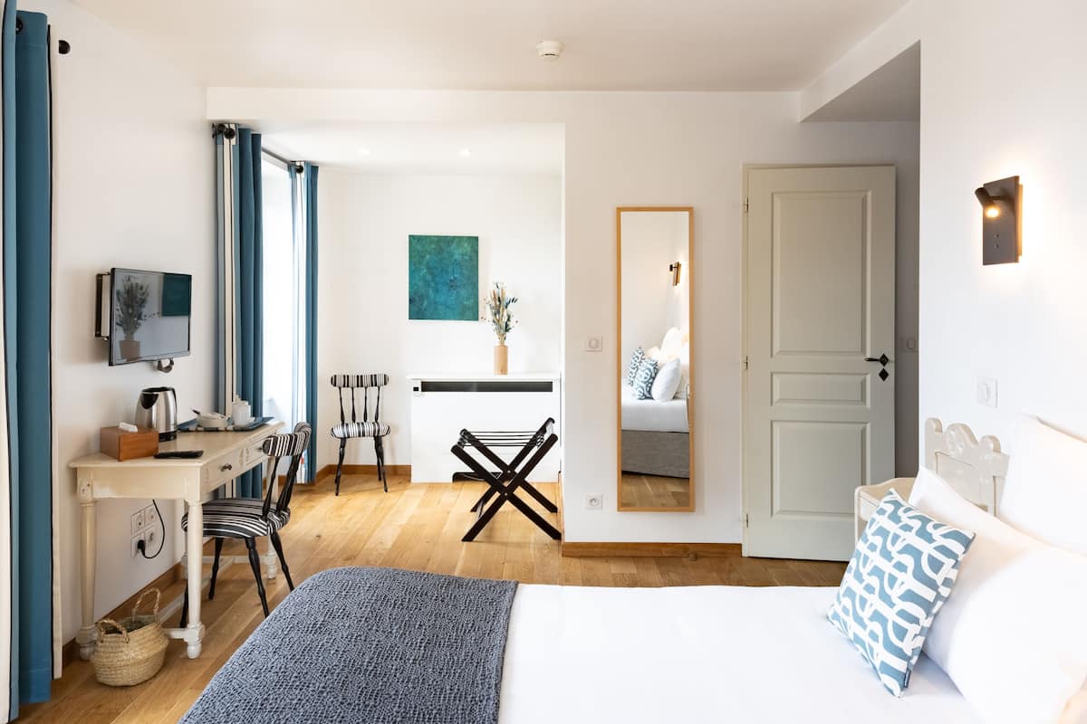 A hotel room with a bed, a desk with a chair, a small table with a plant, a mirror, and a door.