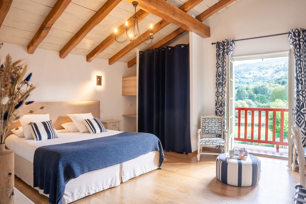 A bedroom with a large bed, a wooden ceiling, a nightstand, a chair, and a balcony with a view of greenery.
