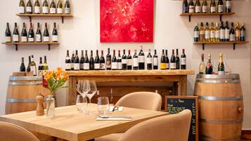 Interior de restaurante con mesa de madera, sillas, botellas de vino en estanterías, un cuadro abstracto rojo y un menú en pizarra.