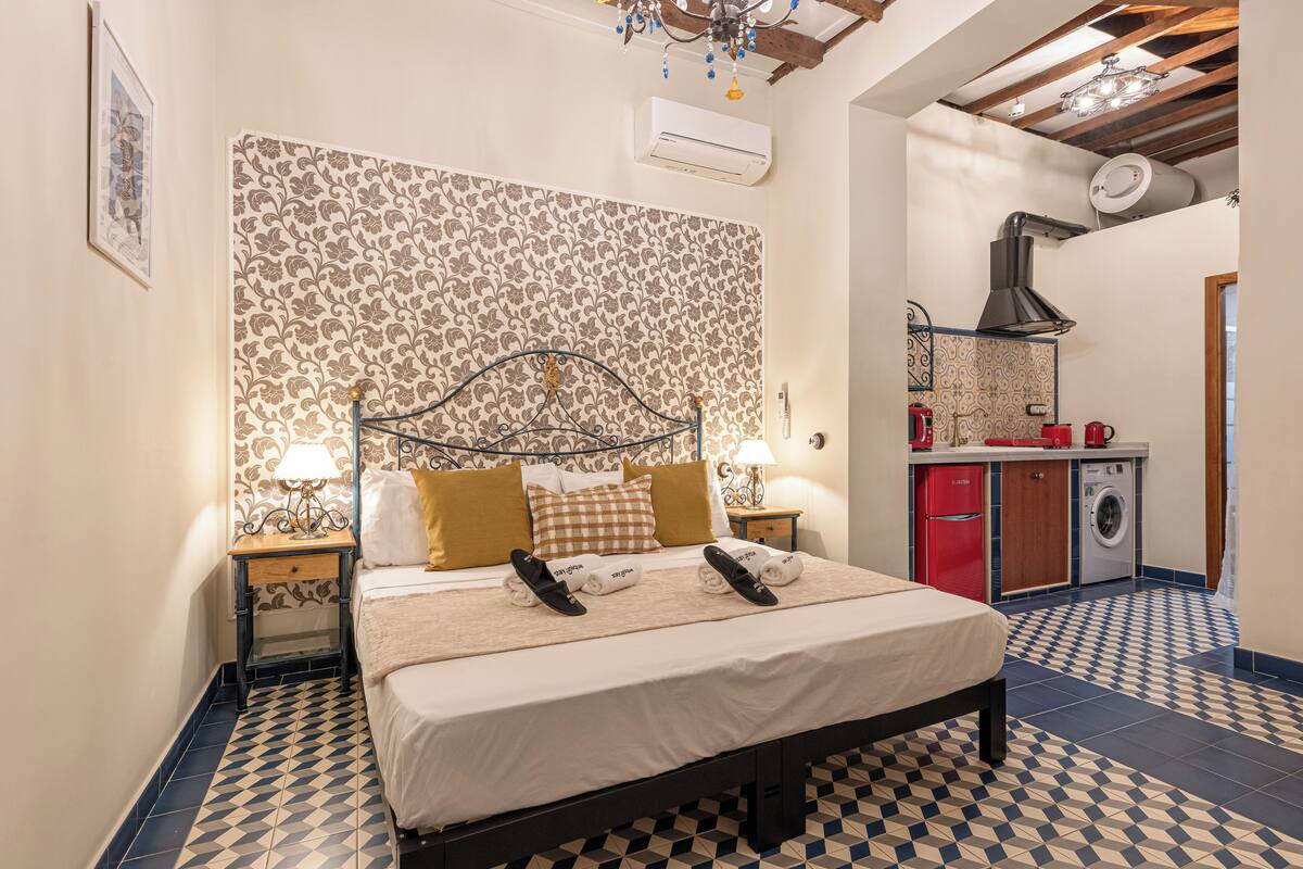 A bedroom with a patterned wallpaper, a bed with a headboard, a nightstand with a lamp, and a small kitchenette with a washing machine.