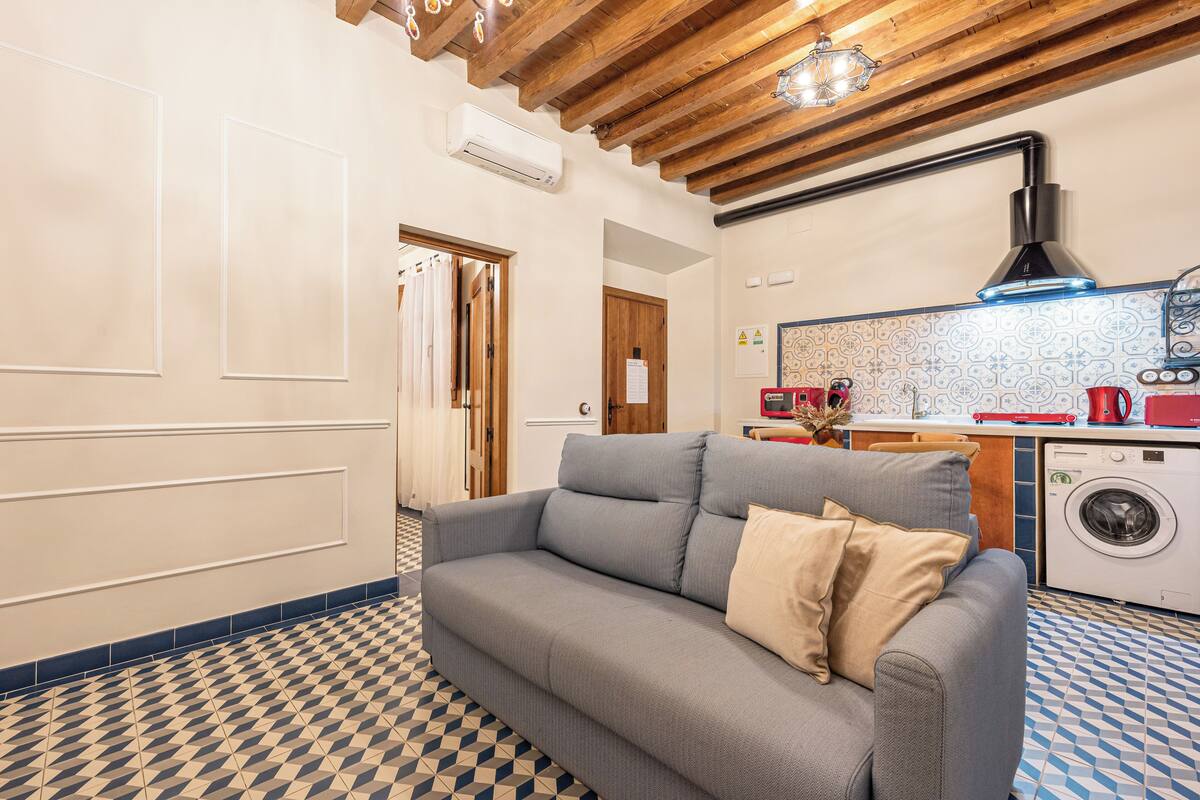 A modern living room with a grey sofa, a washing machine, and a kitchen area with a tiled backsplash.