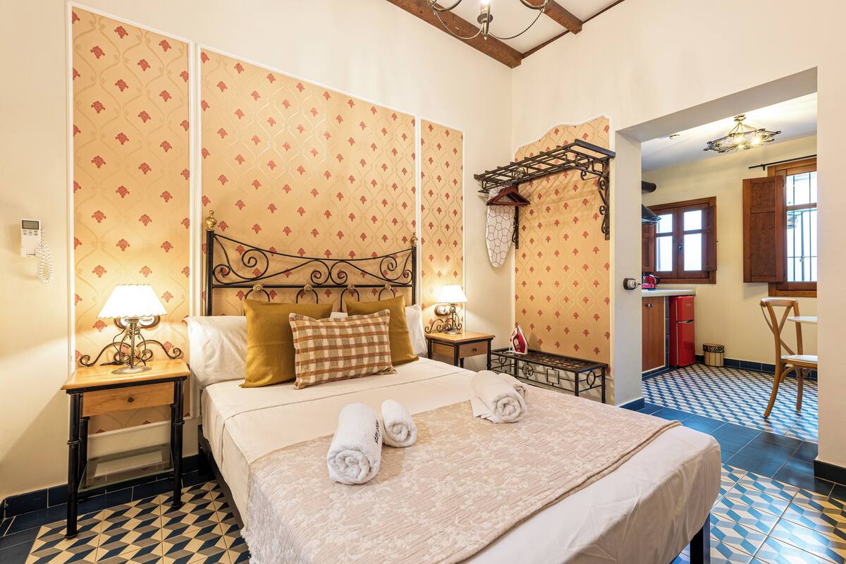 A neatly arranged hotel room with a bed, two bedside tables, a lamp, and a small kitchen area.