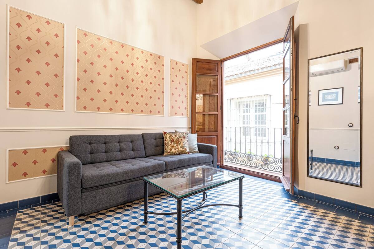A room with a checkered floor, a grey sofa, a glass coffee table, patterned wallpaper, and a door leading to an outdoor balcony.