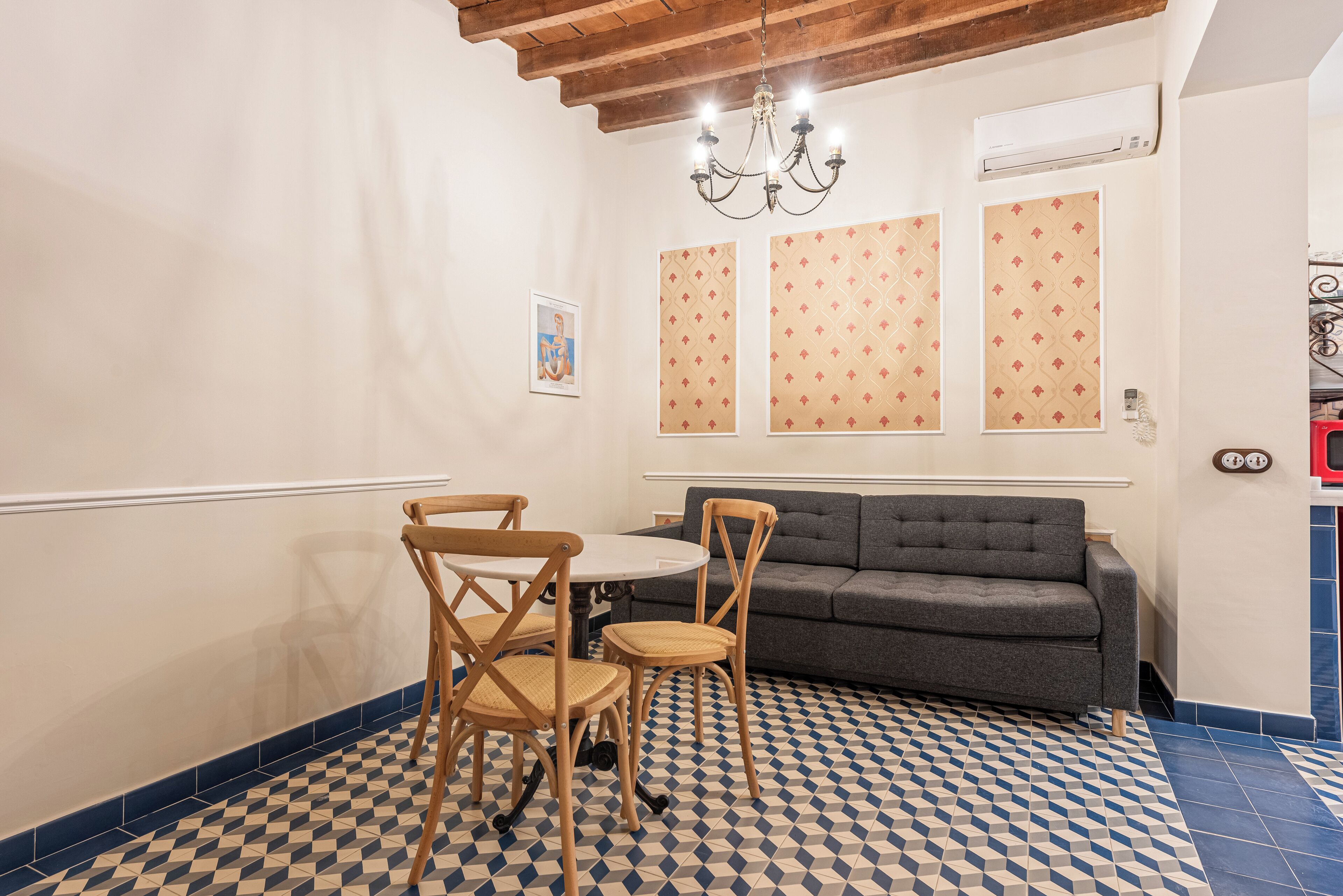 Un comedor con suelo de cuadros, una mesa redonda y dos sillas. Un sofá gris está apoyado contra la pared, y un candelabro cuelga encima.