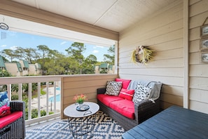 En overdækket balkon med en fletsofa, et lille bord med blomster og udsigt til en swimmingpool og grøntområde.