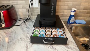 A Keurig coffee maker with a water reservoir and a box of assorted coffee pods on a kitchen counter.