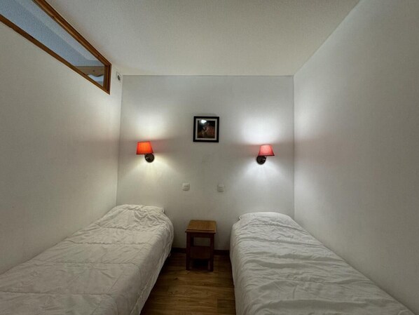 A small, single room with two single beds, a small wooden table, and two wall-mounted lamps.
