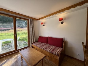 A cozy room with a wooden bench, a small table, and a sofa with red cushions. There are two wall-mounted lamps and a large window with a view of a grassy area.