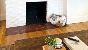 Ein Wohnzimmer mit Kamin aus weißen Ziegelsteinen, einem flachbildfernseher darüber, einem Couchtisch aus Holz mit Büchern und einer Fernbedienung, und einem gemusterten Teppich.