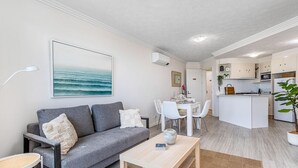 A modern living room with a grey sofa, a wooden coffee table, and a dining area with white chairs. A kitchen with white cabinets and a small island is visible in the background. - Surf Watch Palm Beach (Palm Beach)