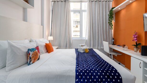 A hotel room with a bed, white walls, an orange accent wall, a desk, and a window with curtains. - Studio in Copacabana 5 Minutes From the Beach Nsc309 (Rio de Janeiro)