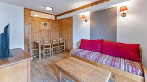 A compact room with a wooden dining table and chairs, a sofa with red cushions, and two wall-mounted lamps.