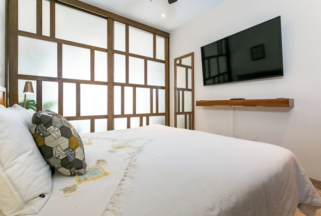 Un dormitorio con una cama, un televisor montado en la pared y una ventana con marcos de madera.