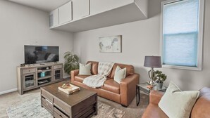 Un salon avec un canapé en cuir marron, une table basse, un téléviseur posé sur un meuble et une fenêtre avec des stores.