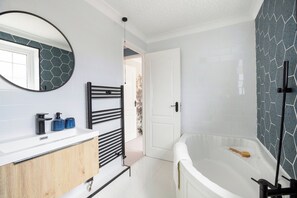 Une salle de bain moderne avec un meuble-lavabo blanc, un porte-serviettes noir, un miroir rond, une baignoire et un espace douche avec un mur à motifs.