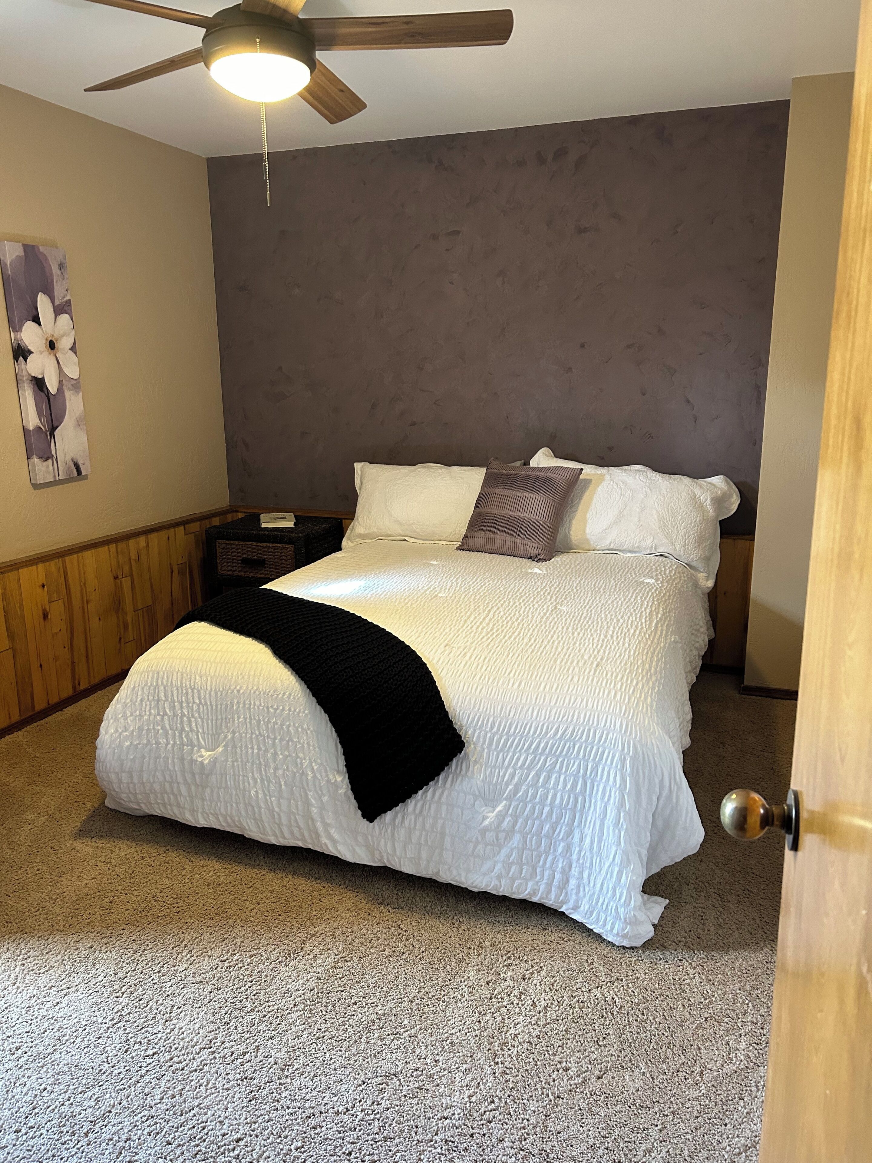 Ein Schlafzimmer mit Bett, Deckenventilator, Nachttisch, Wandbild und einer Tür.
