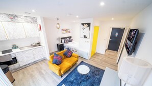 A modern living room with a yellow sofa, a round coffee table, and a blue rug. There is a kitchen area with marble cabinets and a sink.