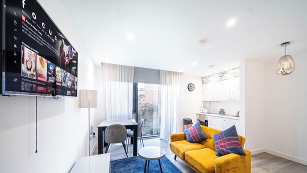 A modern living room with a yellow sofa, a blue rug, a flat-screen TV mounted on the wall, and a dining area with a table and chairs.