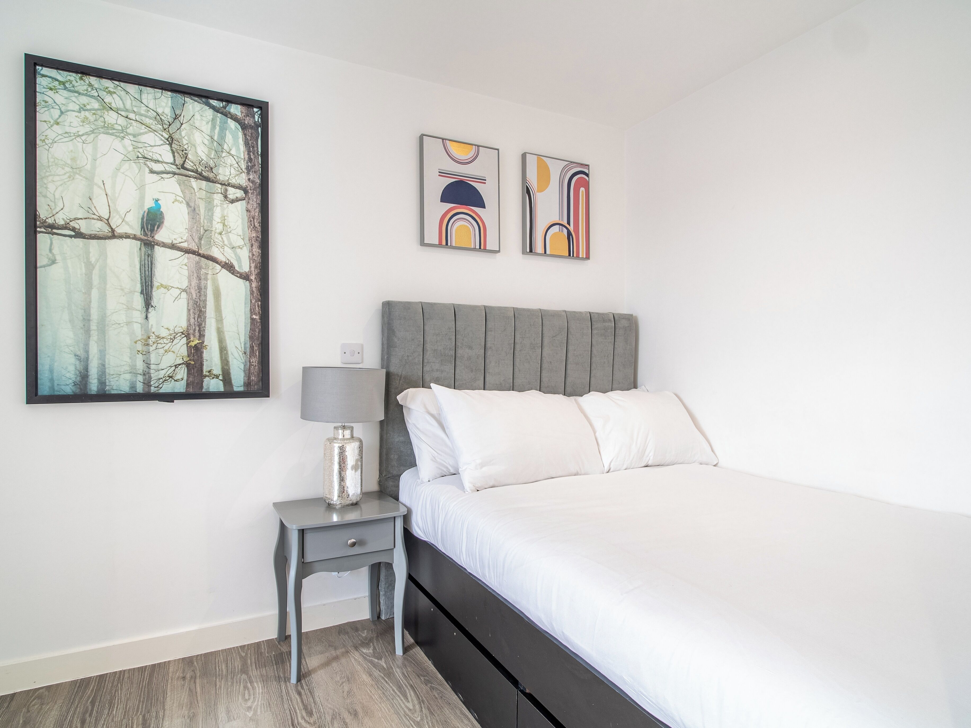 Ein Schlafzimmer mit einem Bett, einem Nachttisch, einer Lampe und zwei abstrakten Bildern an der Wand.