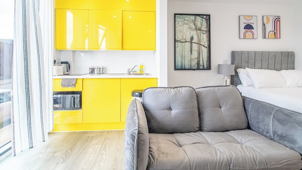 A modern hotel room with a bright yellow kitchen, a grey sofa, a bed with white linens, and artwork on the wall. - Immaculate Studio Apartment in Sheffield City (Sheffield)