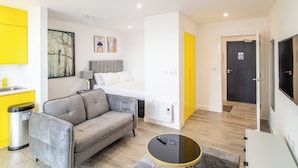 A modern hotel room with a grey sofa, a bed with white bedding, a small table, and a TV. - Immaculate Studio Apartment in Sheffield City (Sheffield)
