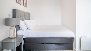 A neatly arranged bedroom with a bed, a bedside table with a lamp, and a wall-mounted power outlet. - Immaculate Studio Apartment in Sheffield City (Sheffield)