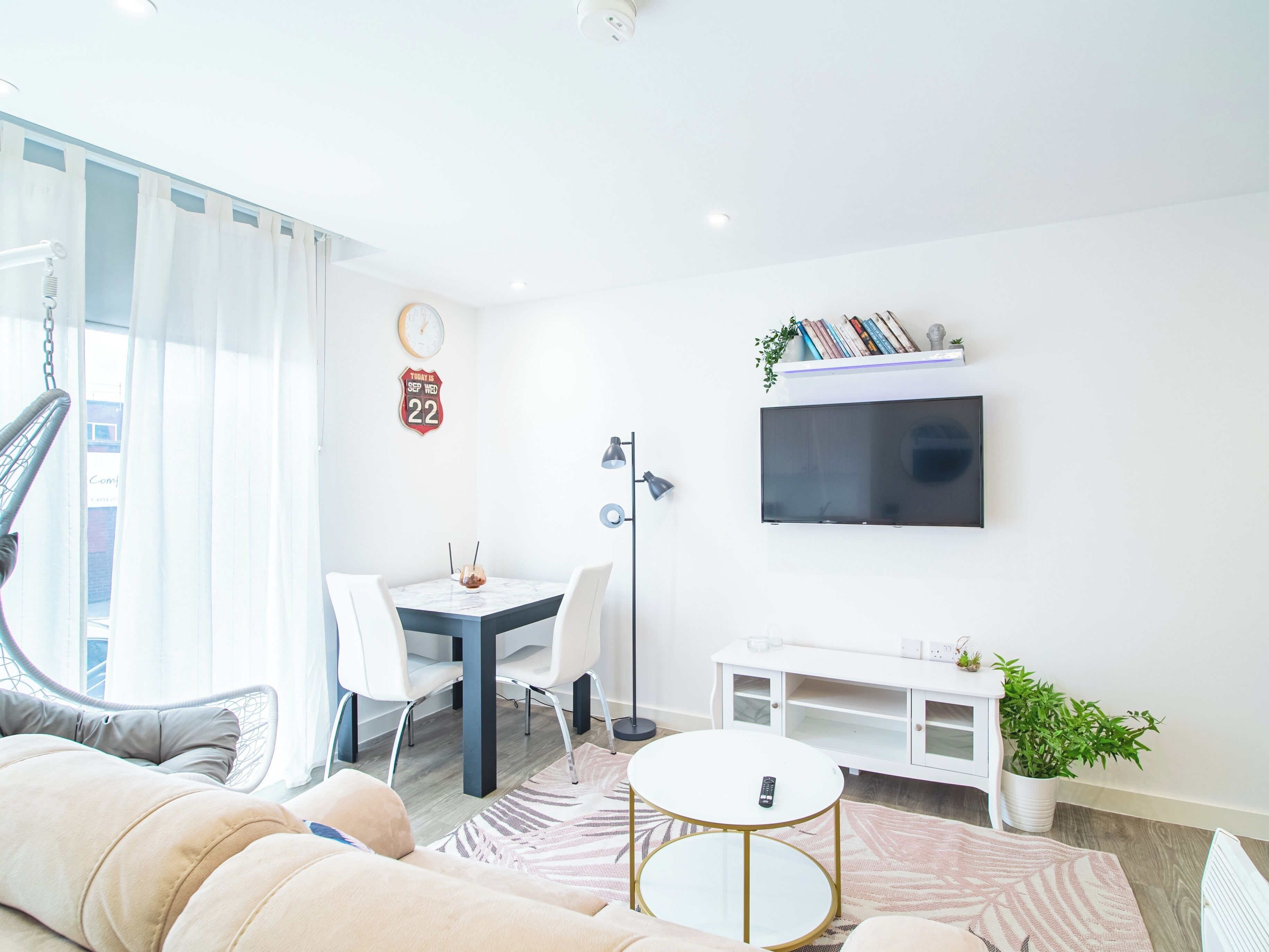 A modern living room with a sofa, a dining table with chairs, a TV mounted on the wall, and a small plant on a white cabinet.