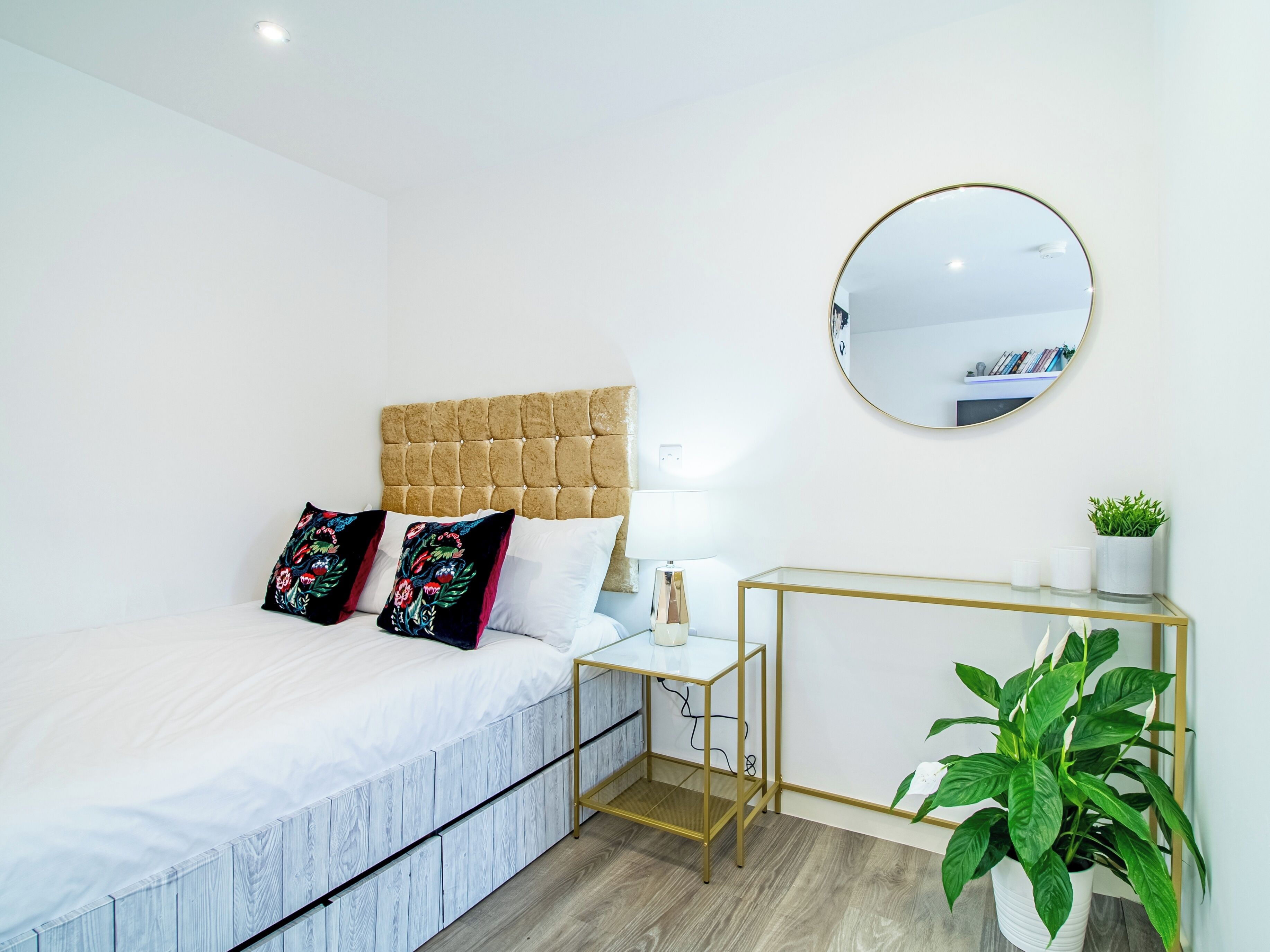A neatly arranged bedroom with a tufted headboard, a bedside table with a lamp, a round mirror, and a small plant.