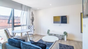 A modern living room with a sofa, armchair, and round tables. A TV is mounted on the wall, and there is a window with curtains. - Deluxe Studio Apartment by Sheffield City Centre (Sheffield)