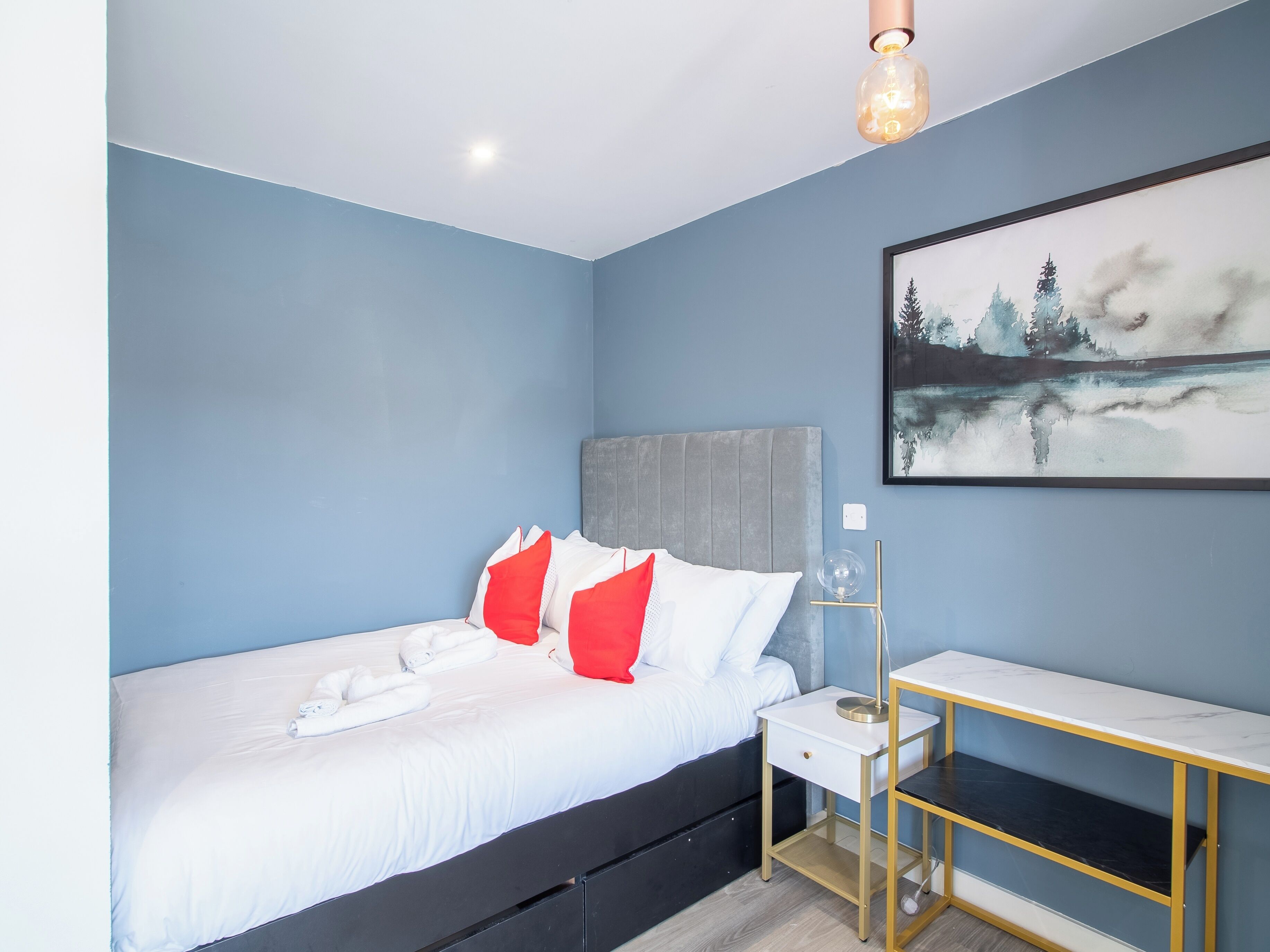 Une chambre à coucher avec un lit, des oreillers, une table de chevet, une lampe et un tableau au mur.