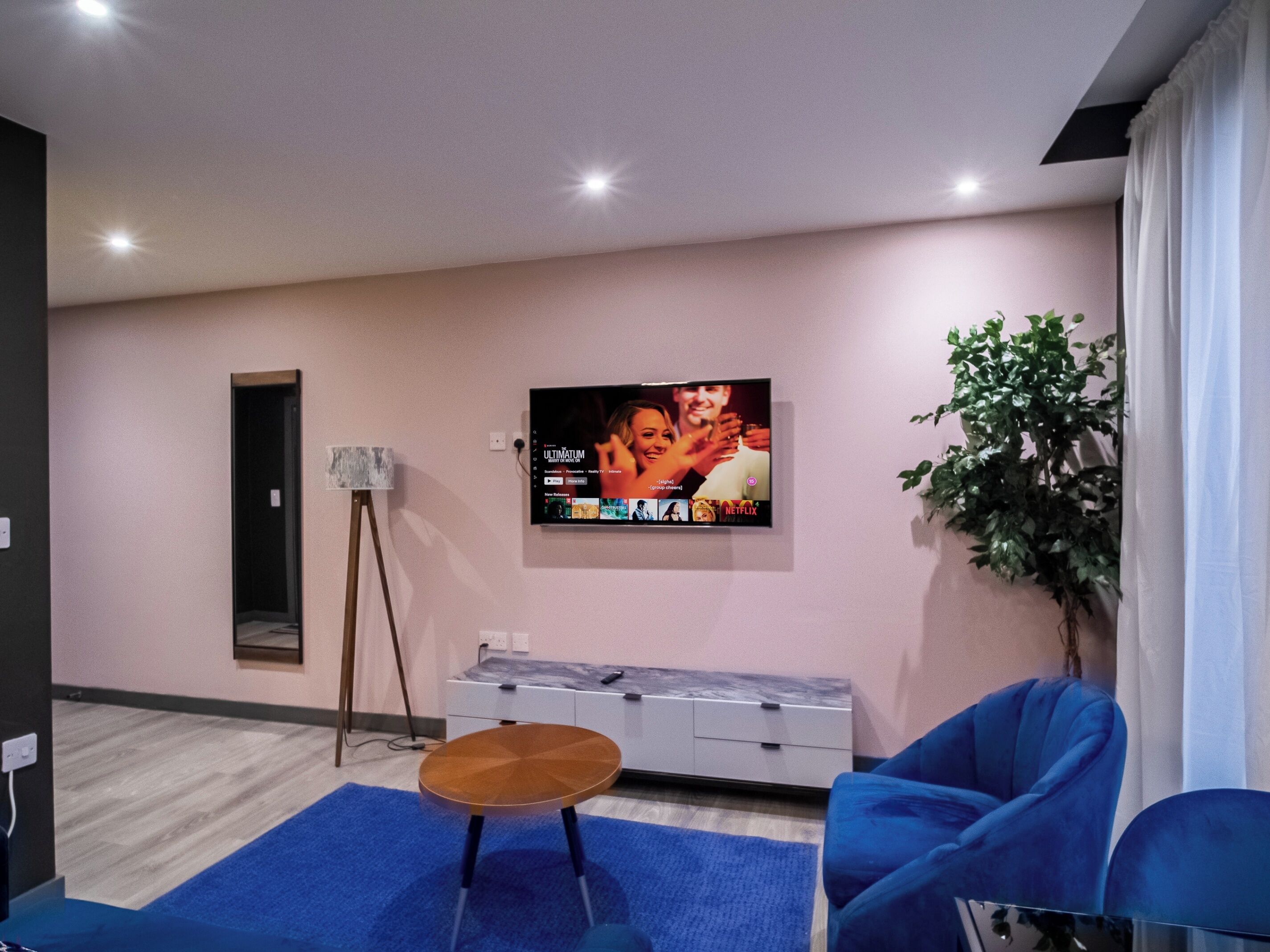Un soggiorno moderno con una televisione a schermo piatto montata al muro, un piccolo tavolino in legno, un divano blu e una pianta in vaso.