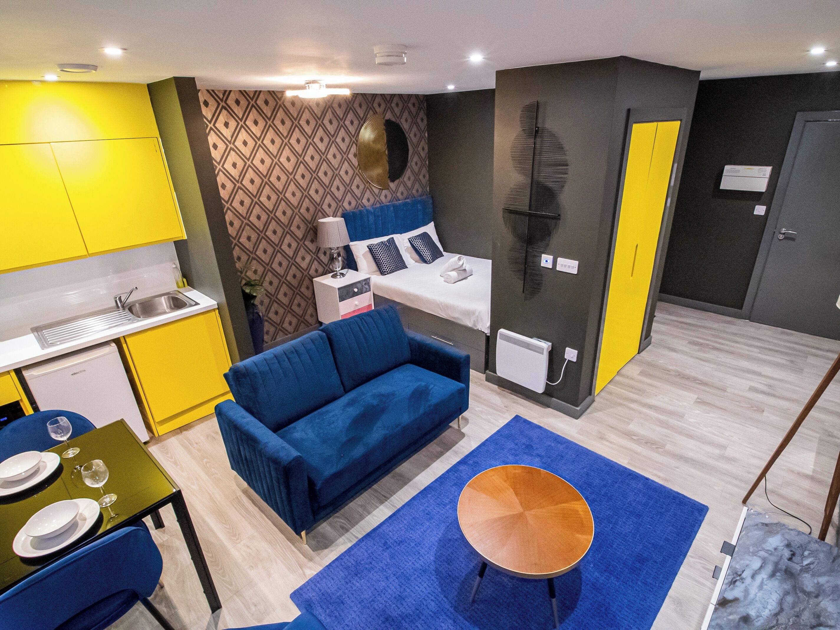A compact living space with a kitchenette, a blue sofa, a small round table, and a patterned wall.