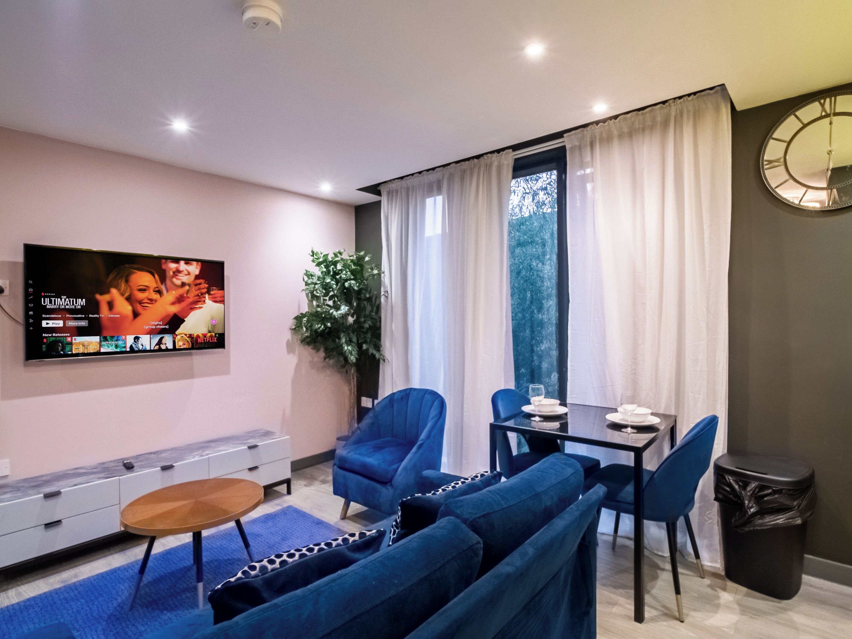 A modern living room with a flat-screen TV, blue sofas, a dining table with chairs, a small wooden side table, and a potted plant.