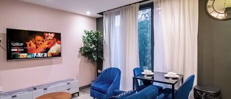 A modern living room with a flat-screen TV, blue sofas, a dining table with chairs, a small wooden side table, and a potted plant.
