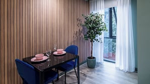 A modern dining area with a wooden panel wall, a dining table with pink plates and blue chairs, a potted plant, and a large window with white curtains. - Central Sheffield City Apartment (Sheffield)