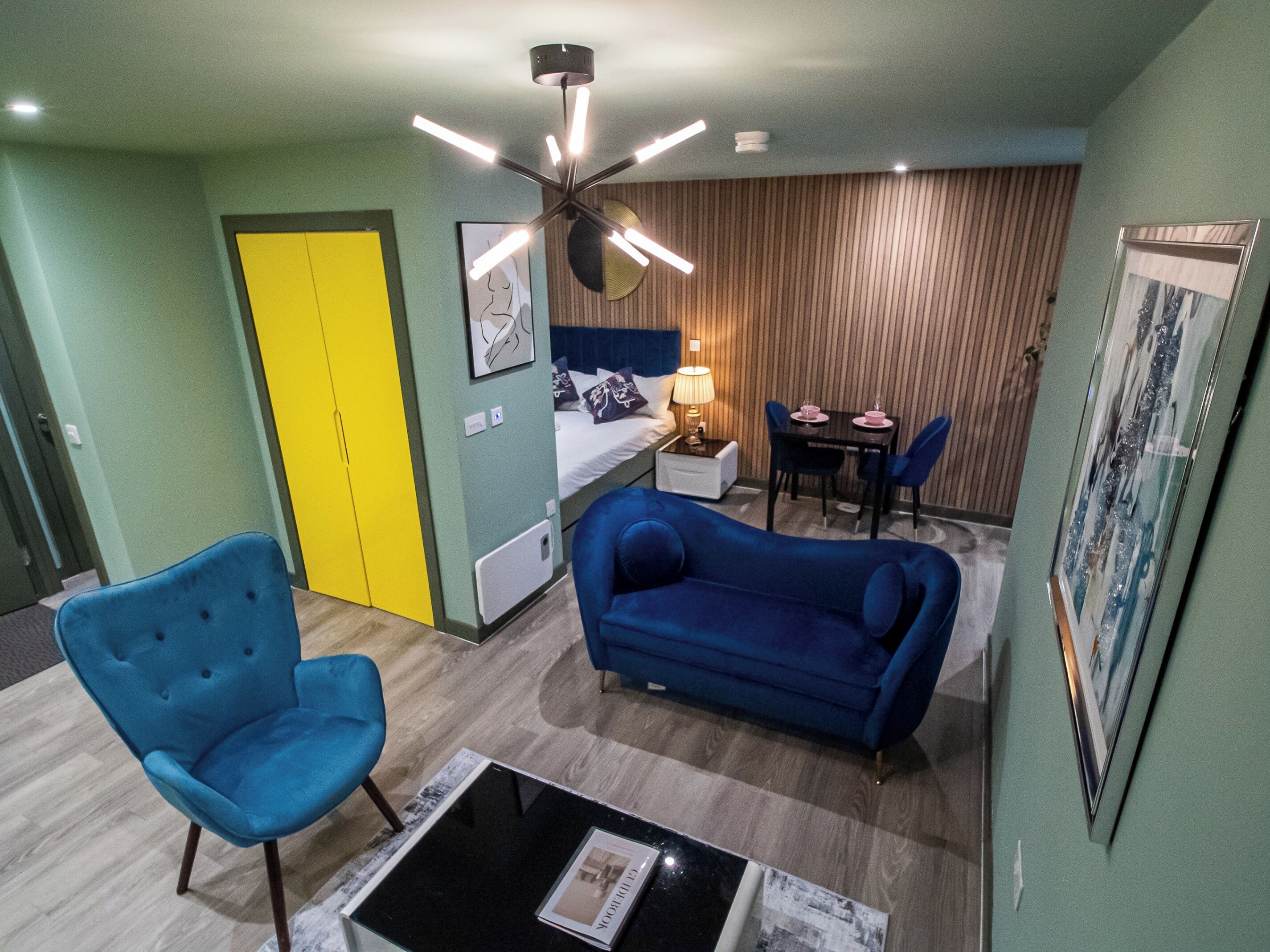 A compact hotel room with a bed, a desk, a chair, a blue sofa, and a small table.