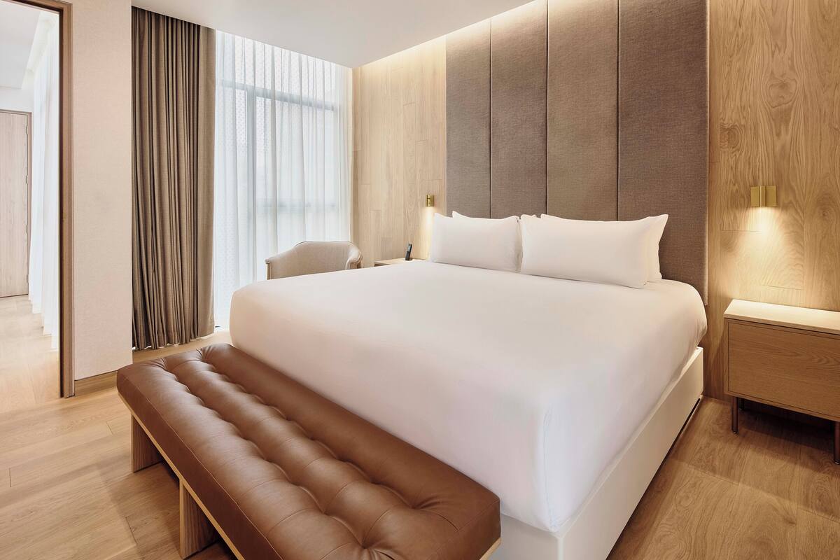 A modern hotel room with a large bed, a wooden headboard, a bench, and a nightstand with a lamp.