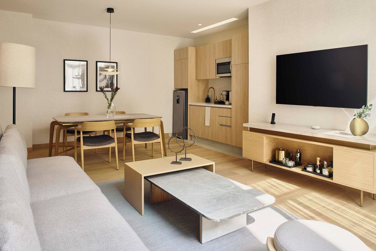 A modern living room with a sofa, coffee table, dining table, and chairs. There is a kitchen area with a refrigerator, microwave, and cabinets. A flat-screen TV is mounted on the wall.