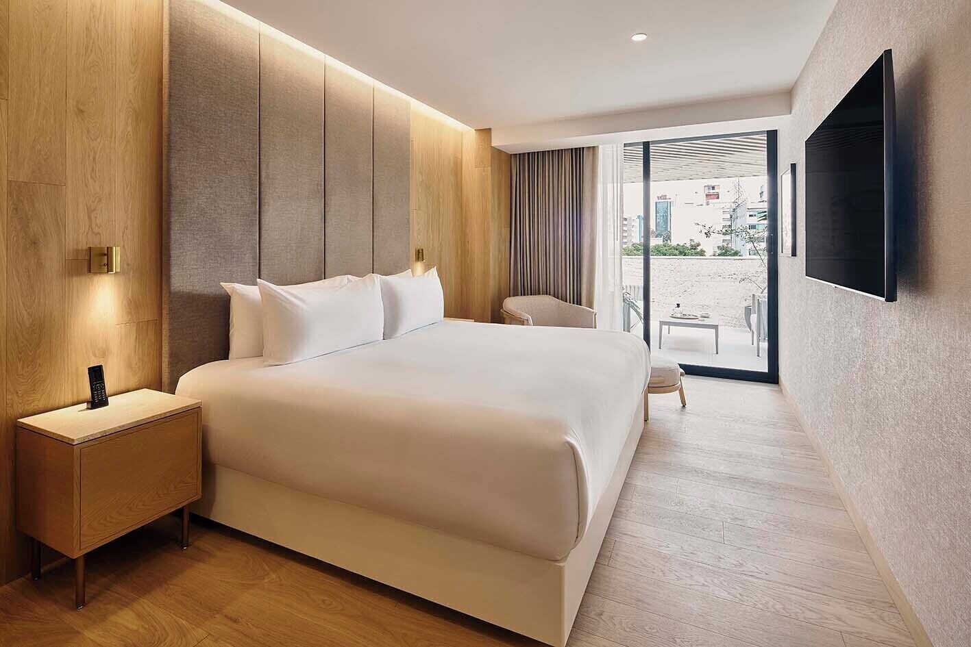 Ein modernes Hotelzimmer mit einem großen Bett, einem Nachttisch mit Fernbedienung, einem Sessel und einem großen Fenster mit Blick auf den Strand.