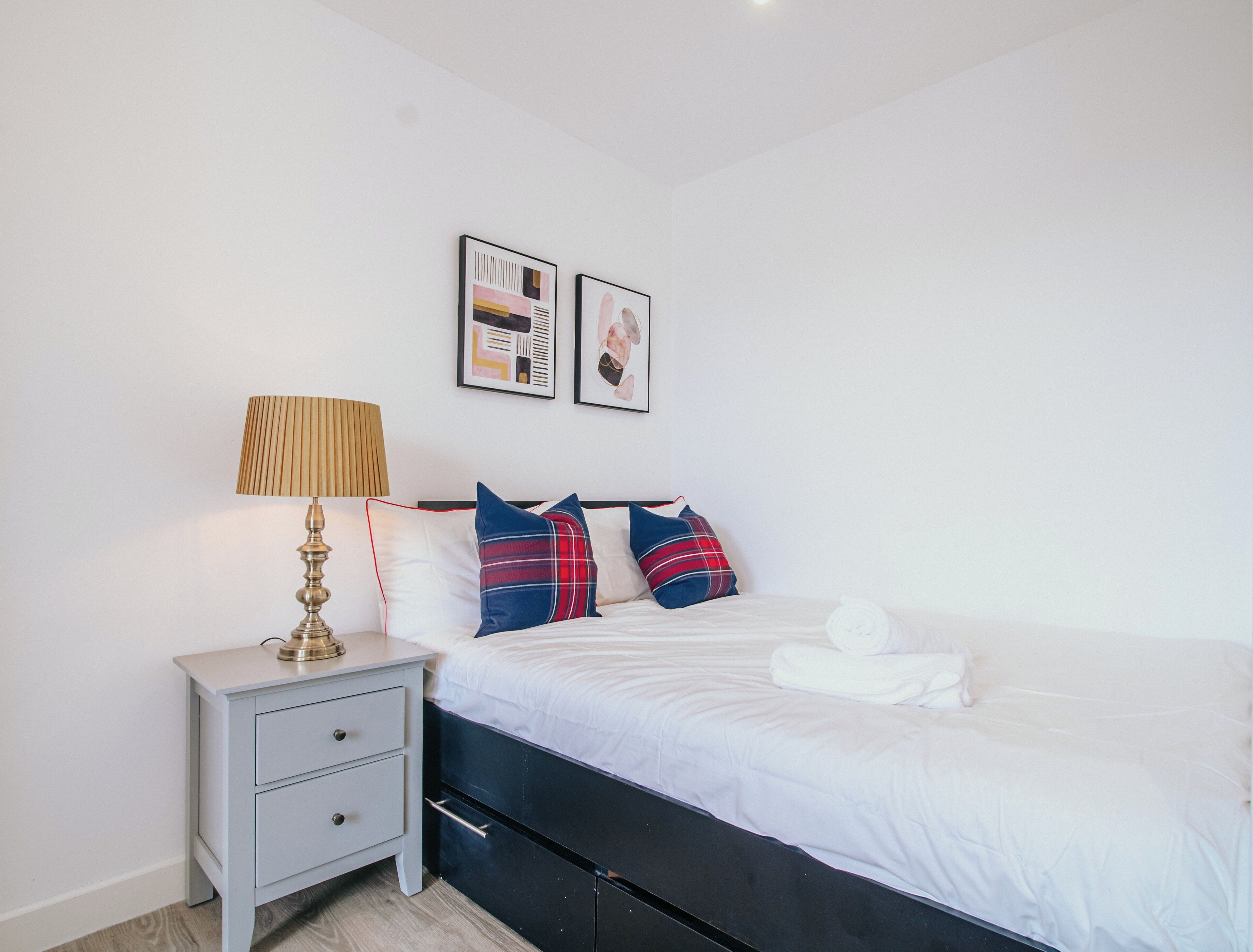 A neatly made bed with white linens, a bedside table with a lamp, and two framed pictures on the wall.