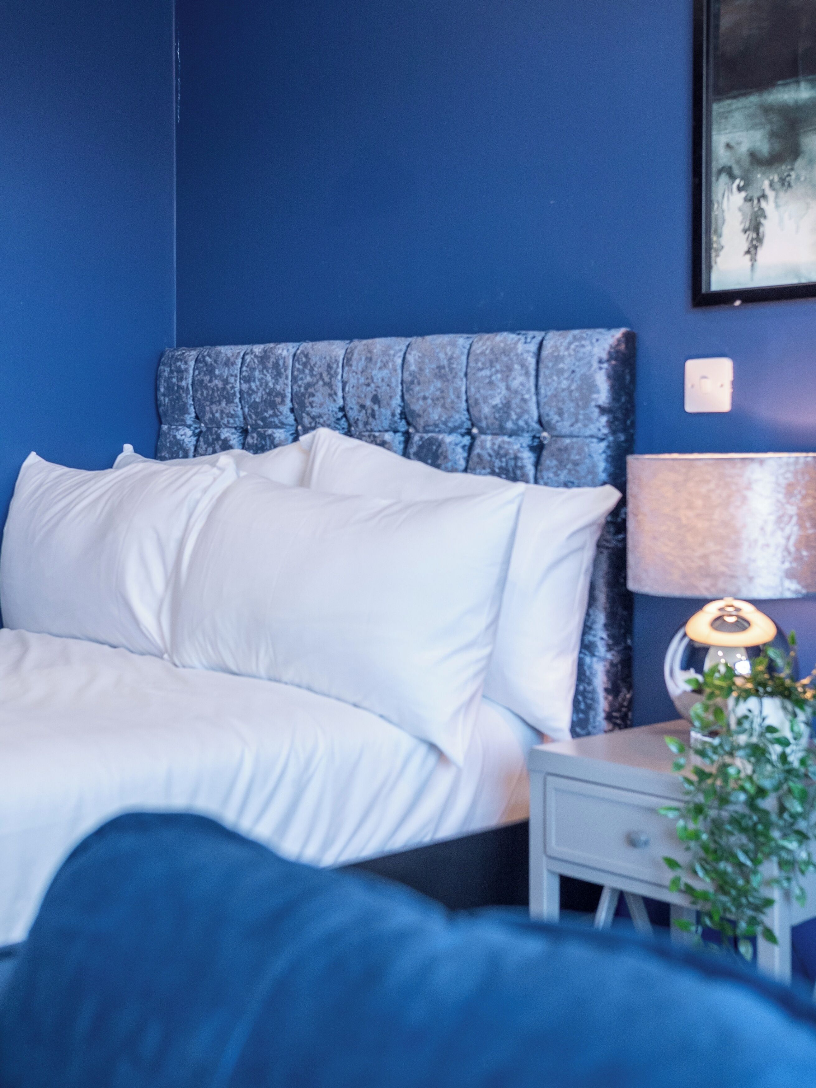 Een bed met witte lakens, een blauw hoofdeinde, een bedlampje en een kleine plant op een nachtkastje.