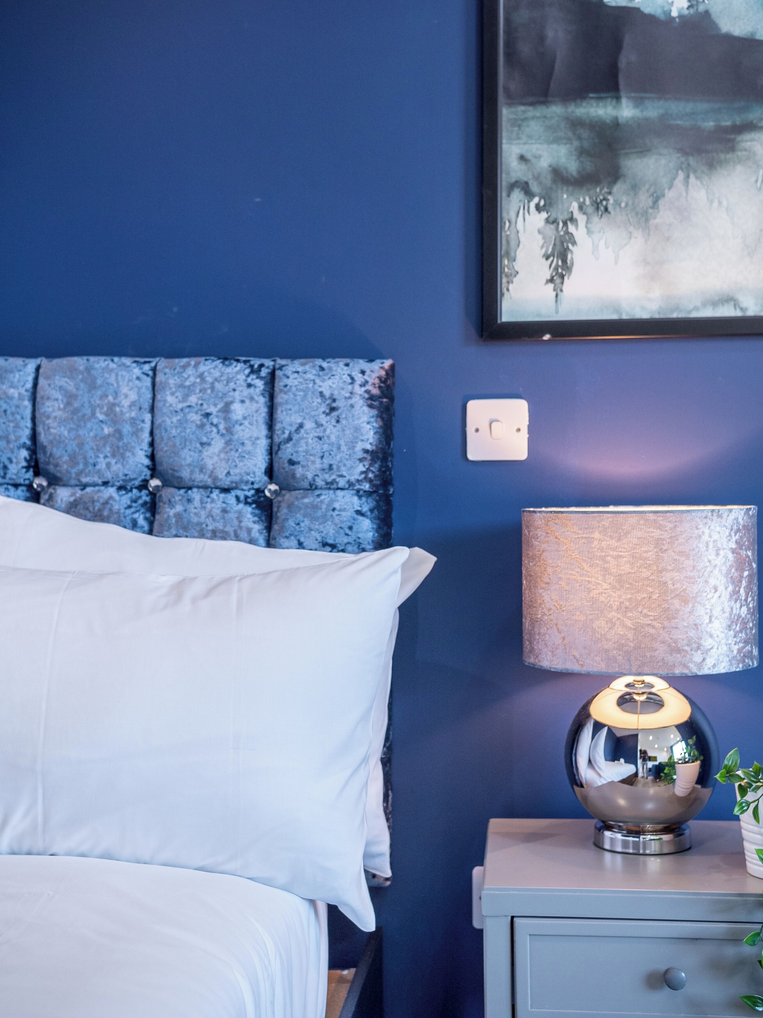 Une chambre à coucher avec une tête de lit capitonnée, un oreiller blanc, une lampe de chevet et une petite table sur laquelle est posée une plante.