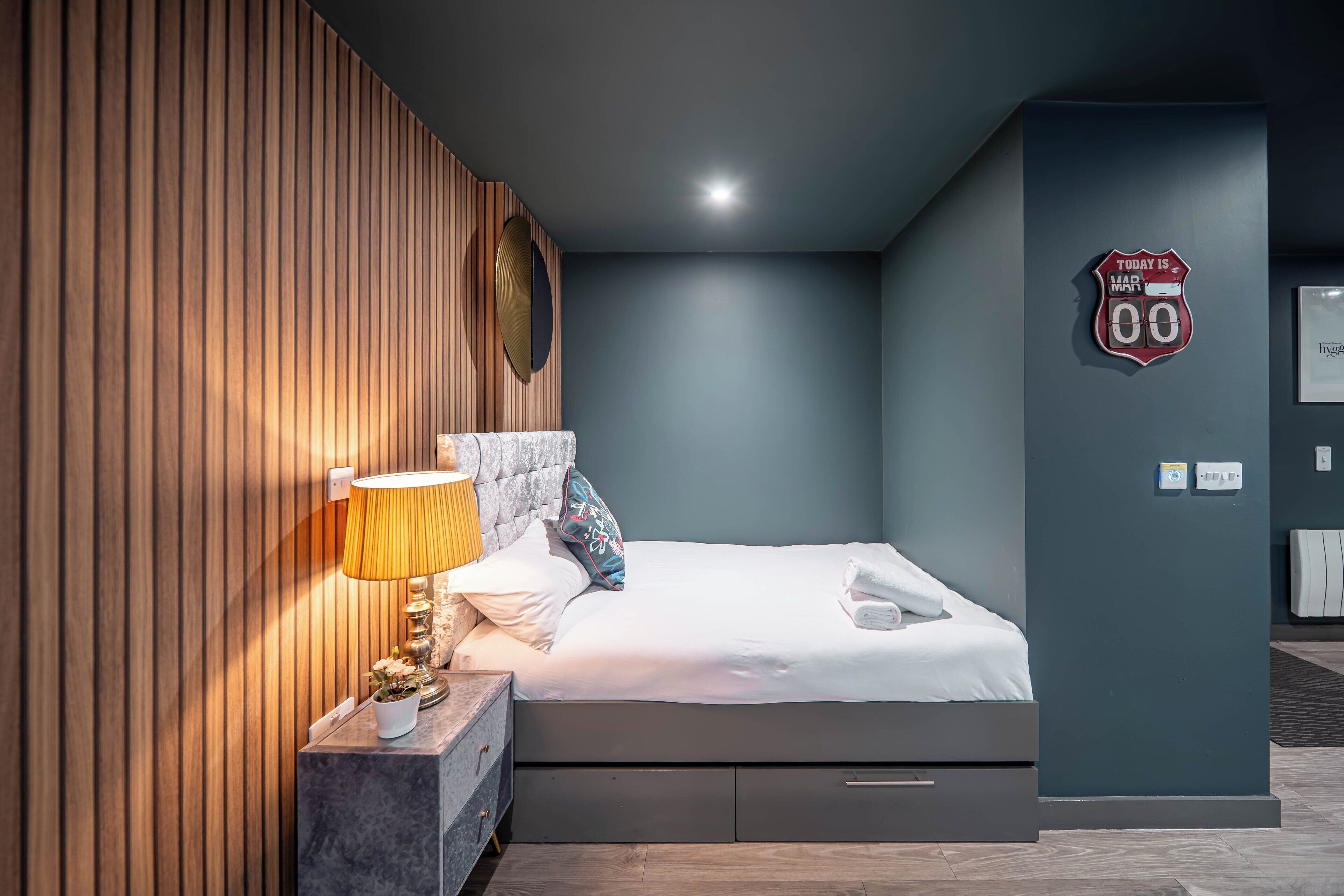 A modern hotel room with a bed, bedside table, lamp, and a wall clock.