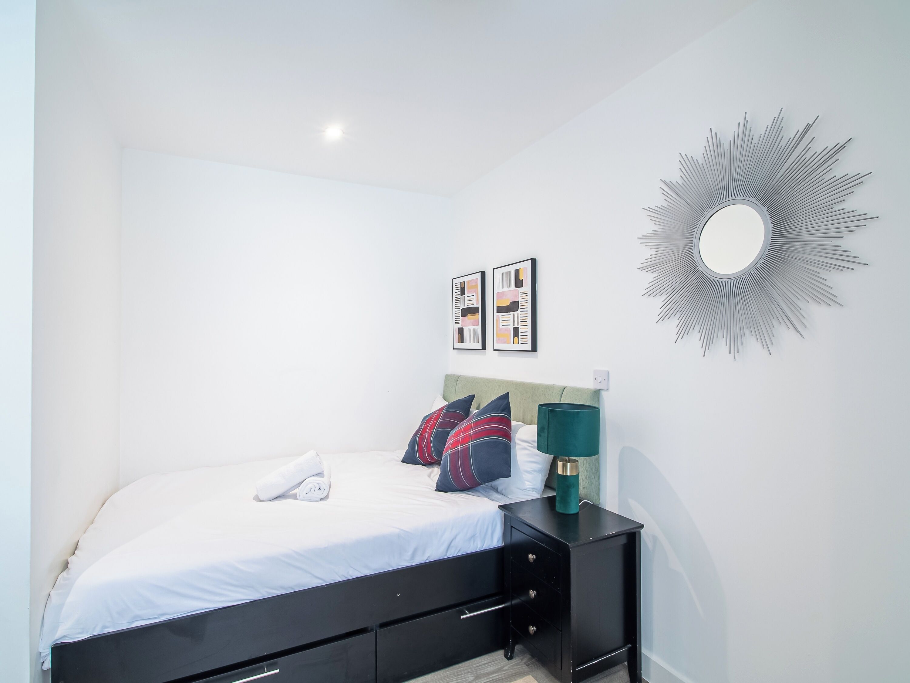 A neatly made bed with white linens and a rolled towel, a black nightstand with a green lamp, and a wall-mounted mirror with a sunburst design.