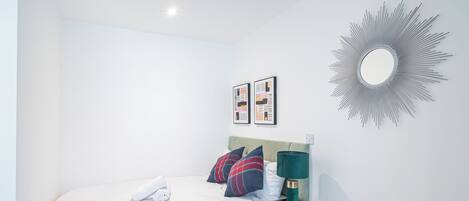 A neatly made bed with white linens and a rolled towel, a black nightstand with a green lamp, and a wall-mounted mirror with a sunburst design.