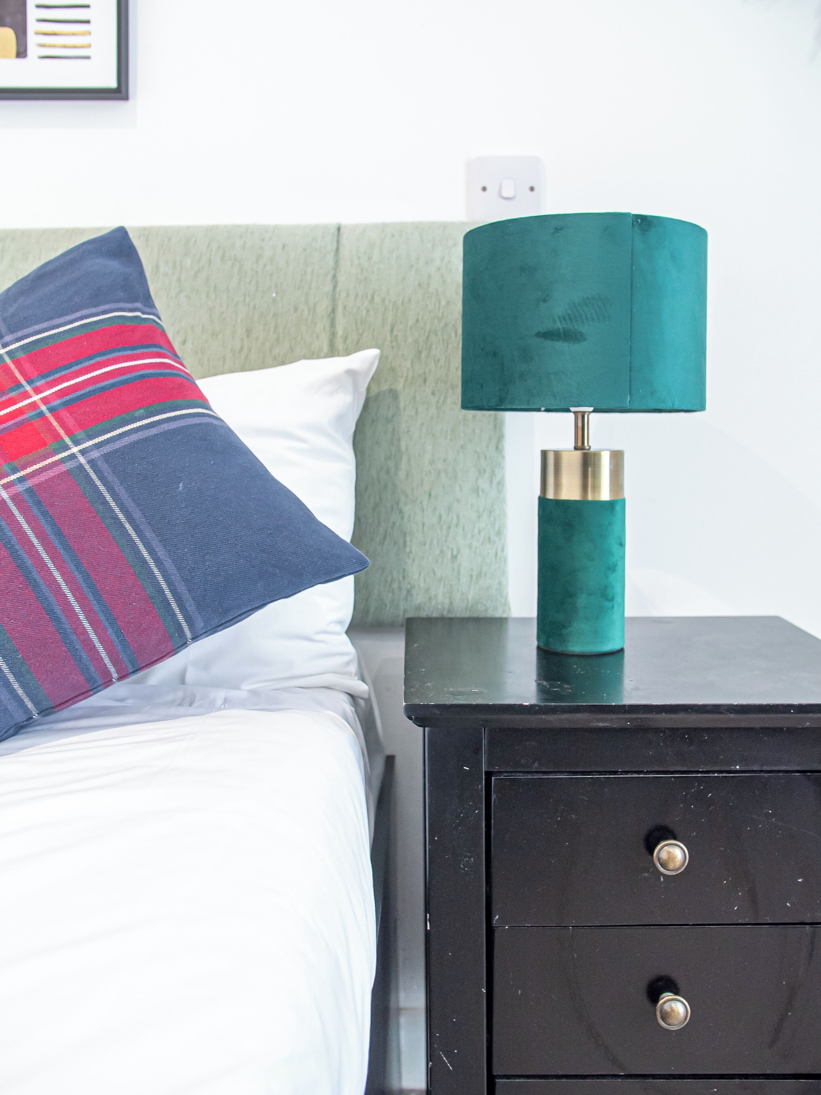 A bed with a plaid pillow and a teal lamp on a black nightstand.