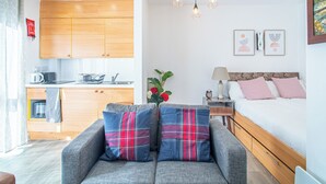 Une chambre moderne avec un canapé gris, un lit en bois, une petite table de chevet et un coin cuisine doté de meubles en bois et d’un four à micro-ondes.